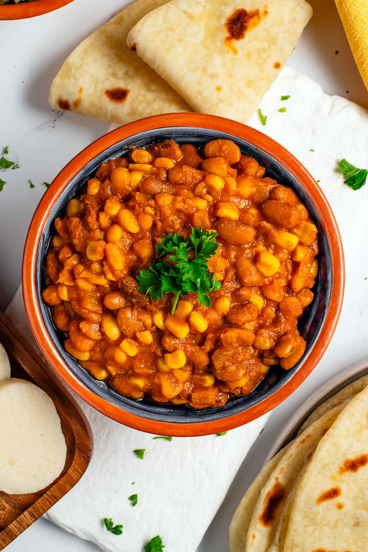 Top down view of Githeri, a Kenyan bean and rice stew. This version has a thick tomato gravy and uses pinto beans and white navy beans. The githeri is garnished with fresh parsley. Soft Kenyan chapati and slices of ugali lay around the bowl of githeri.