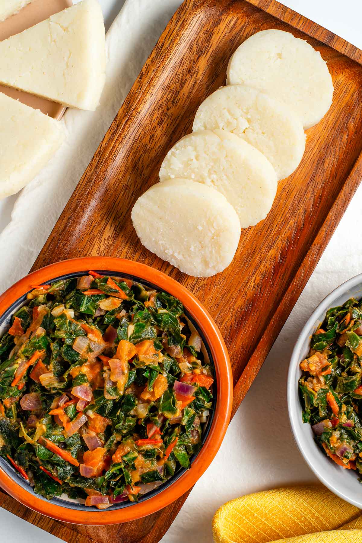 Top down view of round slices of ugali resting on a wooden tray next to a small bowl of sukuma wiki.