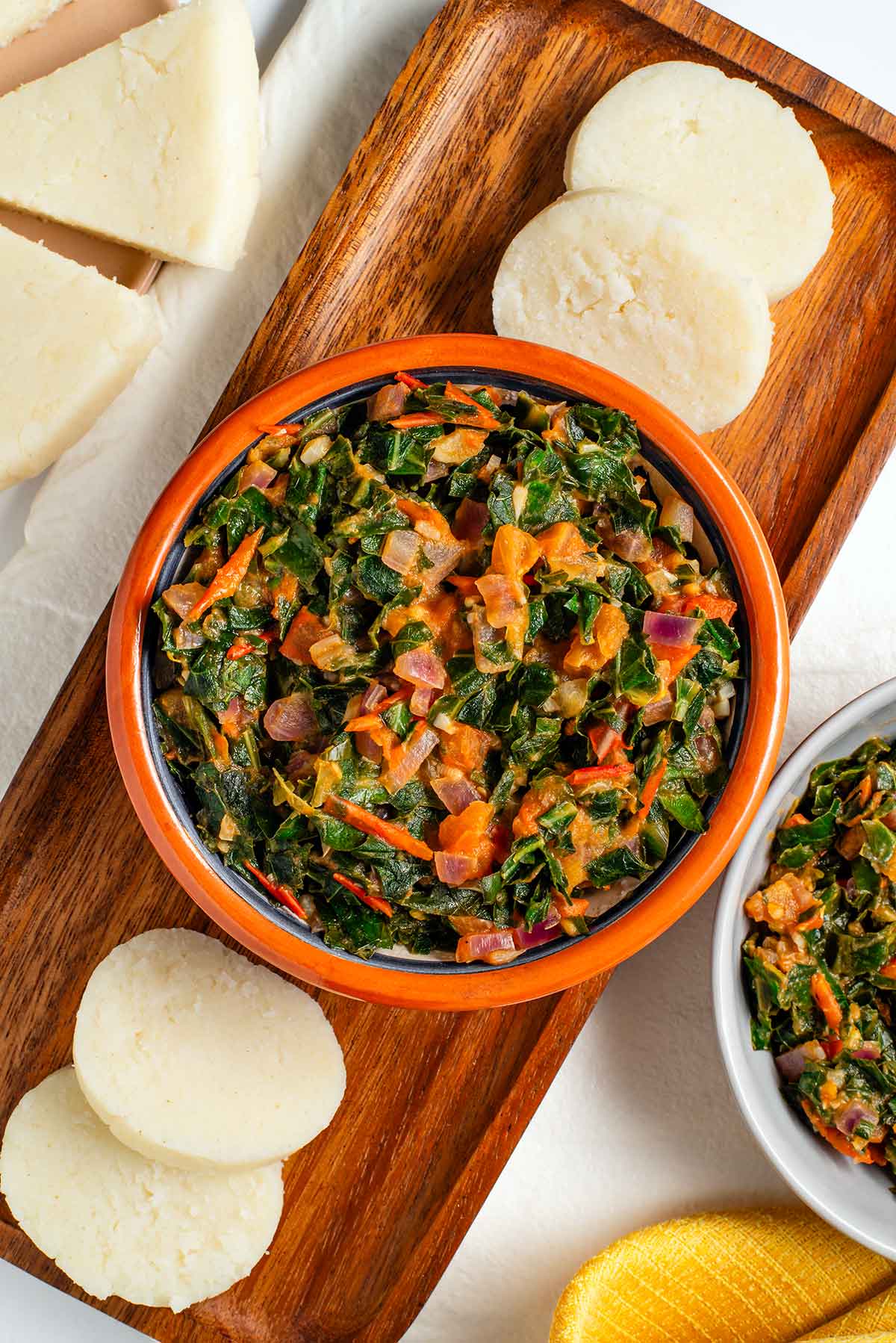 Top down view of a small bowl filled with sukuma wiki. The sauteed collard greens are mixed with garlic, onion, and tomato. Round slices of ugali rest beside the bowl and pie shaped slices of ugali can also be seen.