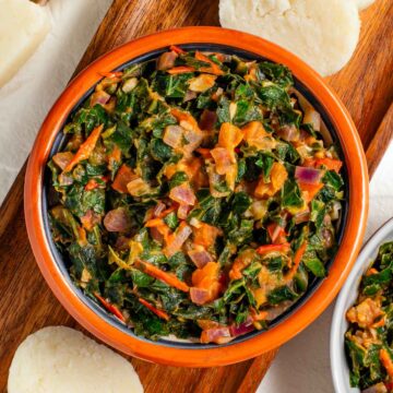 Top down view of a small bowl filled with sukuma wiki. The sautéed collard greens are mixed with garlic, onion, and tomato. Round slices of ugali can be seen resting beside the bowl.