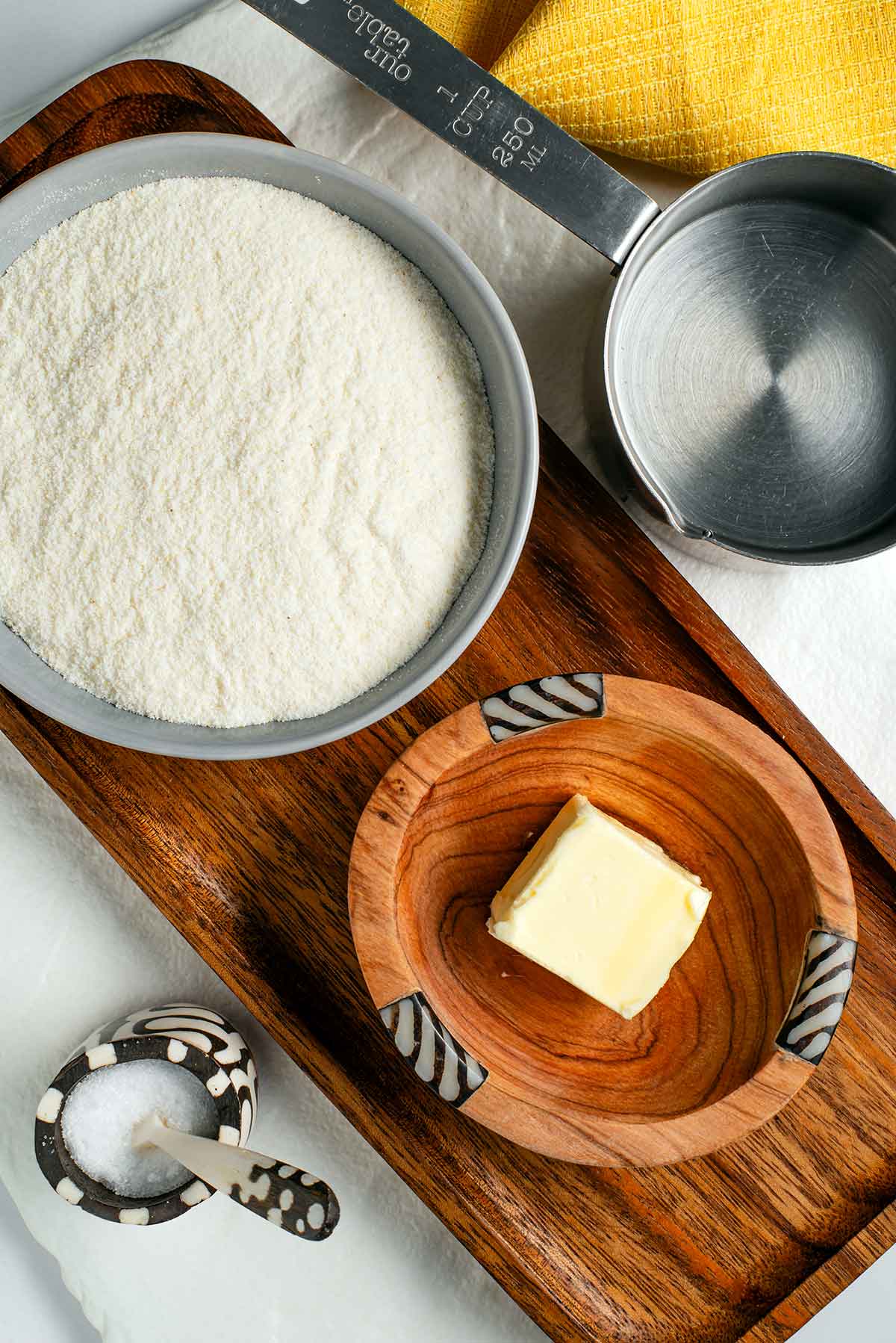 Top down view of ingredients on a wooden tray. A small bowl is filled with fine white cornmeal, a measuring cup holds water, and small Kenyan wooden bowls hold salt and vegan butter.