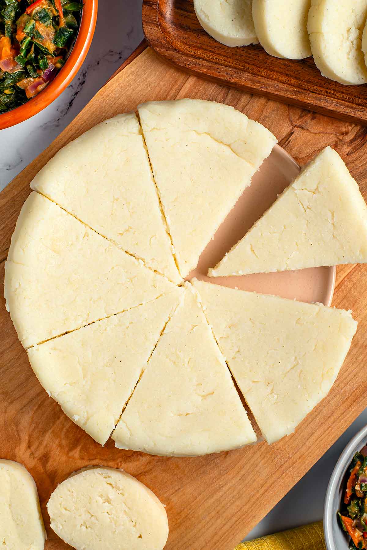 Top down view of pie shaped slices of Kenyan ugali. The cornmeal is firm, smooth, and white. Circular slices of ugali are also shown with sukuma wiki in small bowls at the edges of the frame.