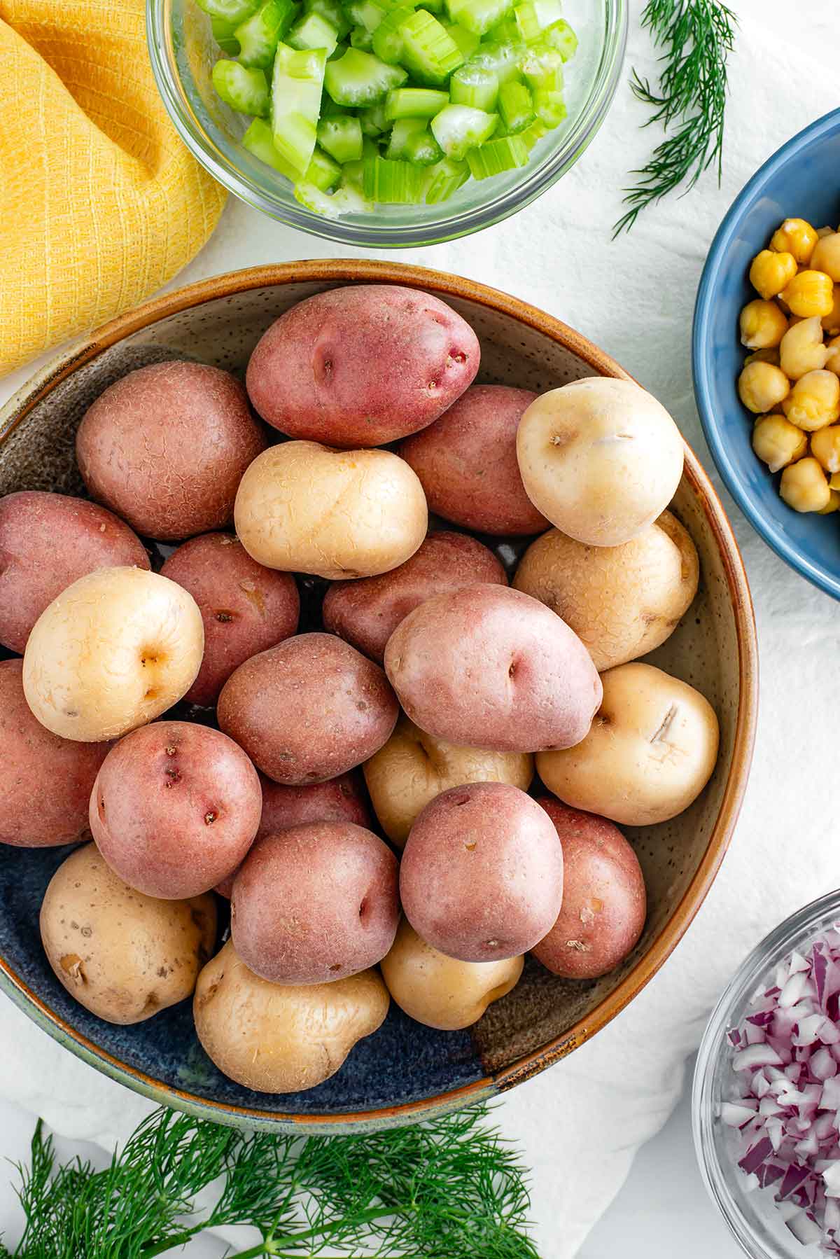 Top down view of baby red and yellow potatoes in a bowl. Sprigs of fresh dill lay on a tray while bowls of celery, chickpeas, and red onion surround the potatoes.
