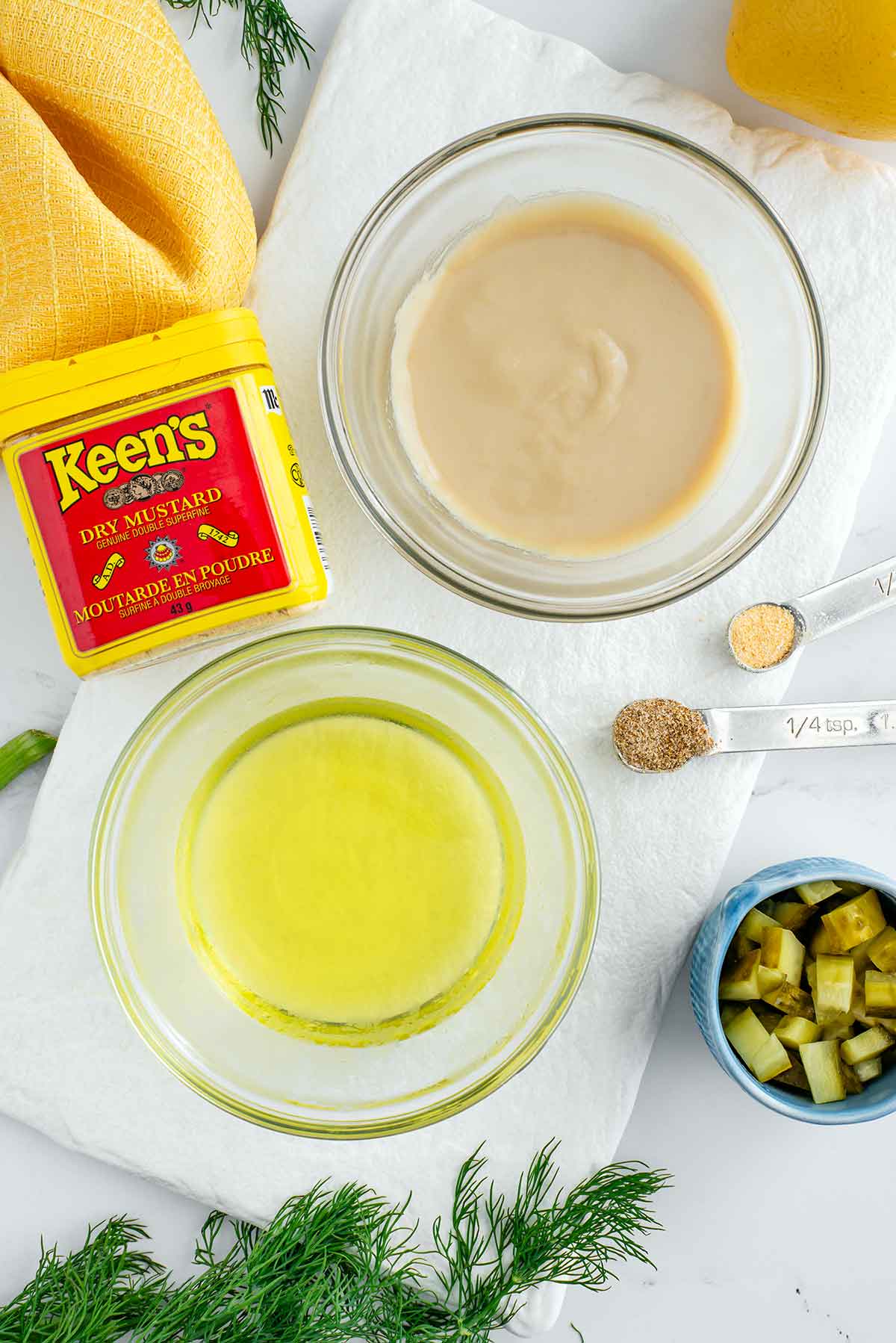 Top down view of two small bowls on a white tray. One holds creamy tahini paste and the other holds pickle juice. Dry mustard powder, a lemon, some other spices, fresh dill, and chopped pickles surround the bowls.