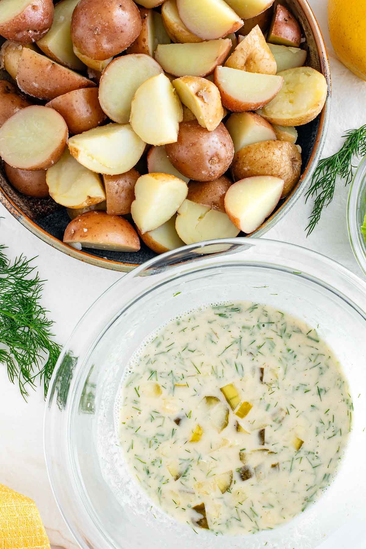 Top down view of quartered and cooked baby potatoes next to a bowl of creamy dressing. The dressing is speckled with fresh chopped dill and chopped pickles.