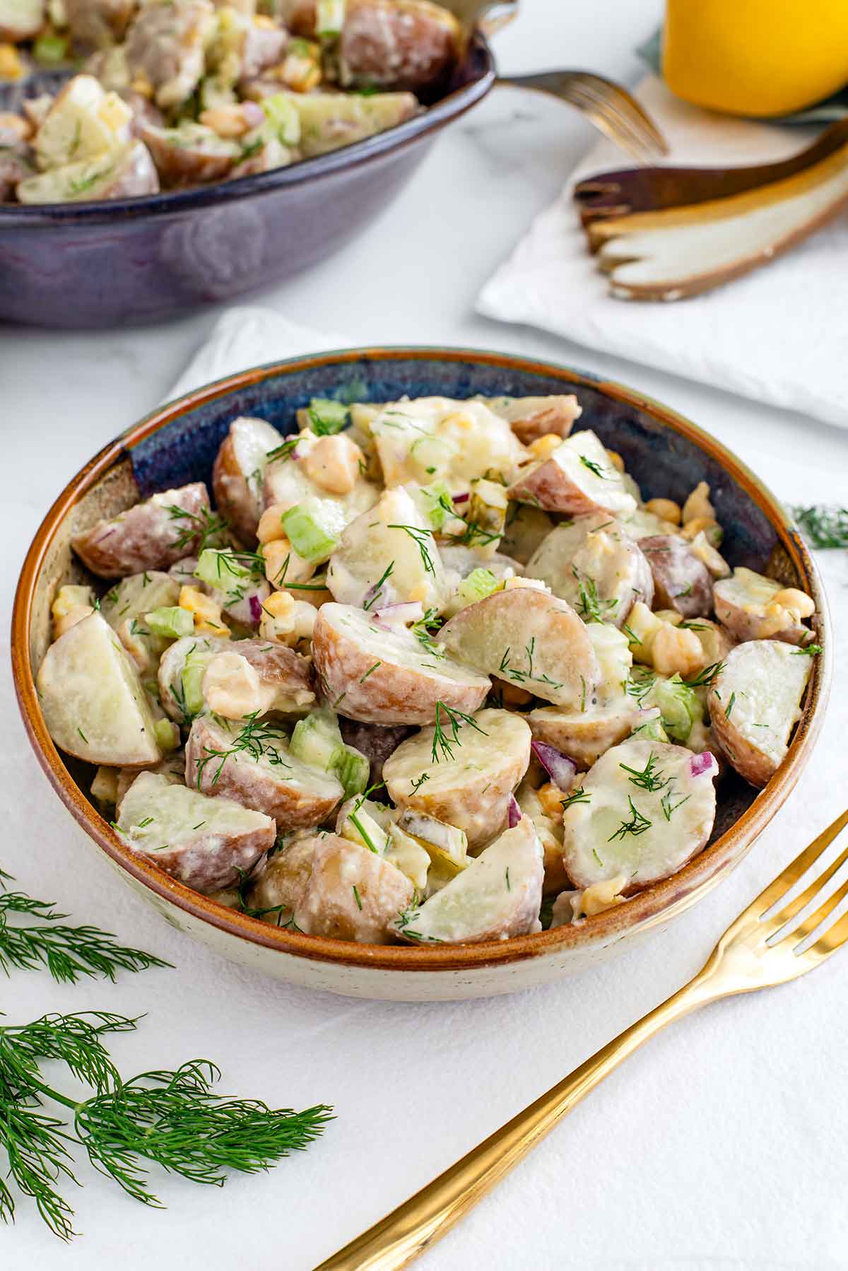 Side view of vegan potato salad in a small serving bowl. The red and yellow skinned potatoes are covered in a creamy dressing and the salad is speckled with fresh dill, chickpeas, red onion, celery, and pickles. Another serving bowl rests in the background.