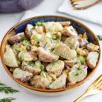 Side view of vegan potato salad in a small serving bowl. The red and yellow skinned potatoes are covered in a creamy dressing and the salad is speckled with fresh dill, chickpeas, red onion, celery, and pickles.