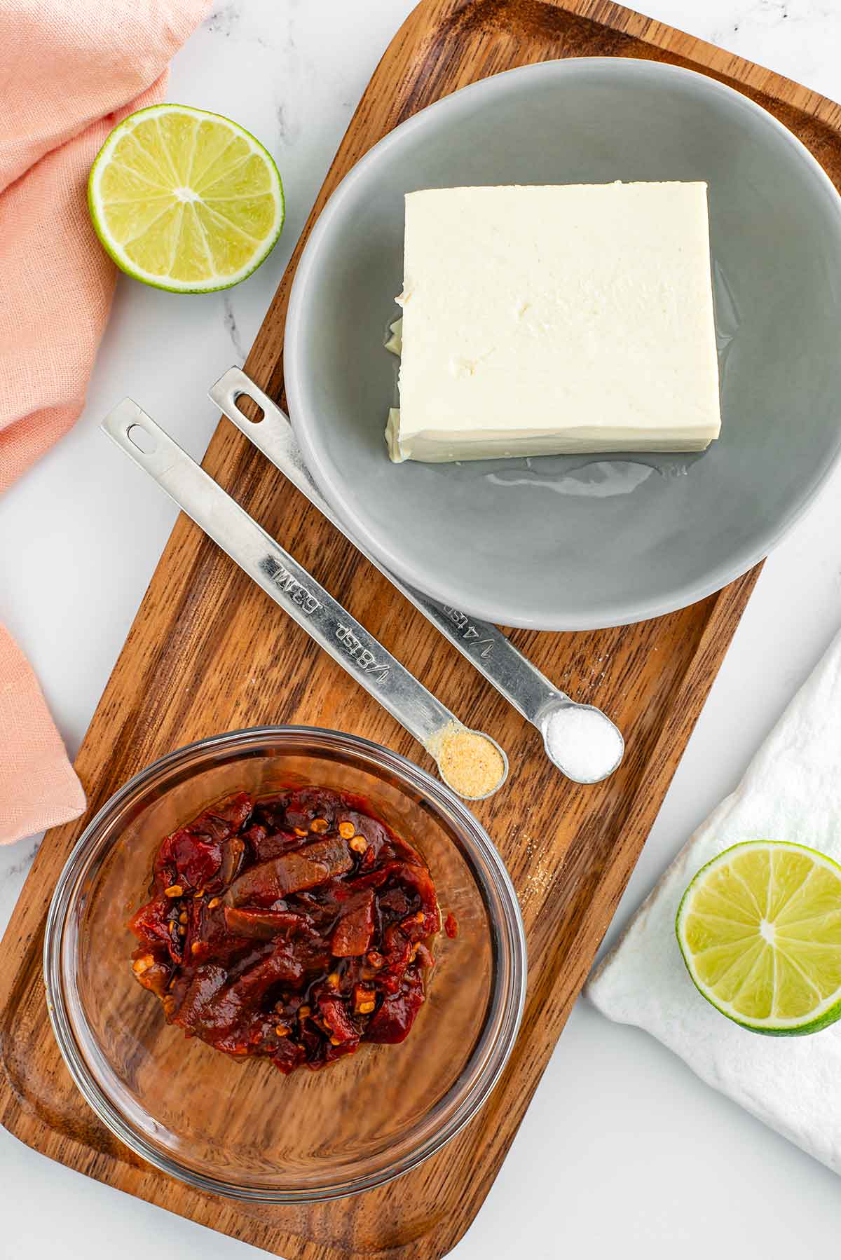 Top down view of ingredients on a wooden tray. Soft tofu rests in a bowl, a small dish contains diced chipotle pepper in adobo sauce, a fresh cut lime is pictured, and two measuring spoons contain salt and garlic powder.