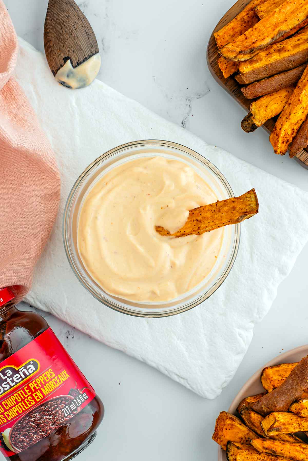 Top down view of blended oil-free chipotle mayo in a small glass bowl. A sweet potato fry rests in the thick mayo and a jar of chipotle peppers in adobo sauce rests on the tray beside the prepared mayo.