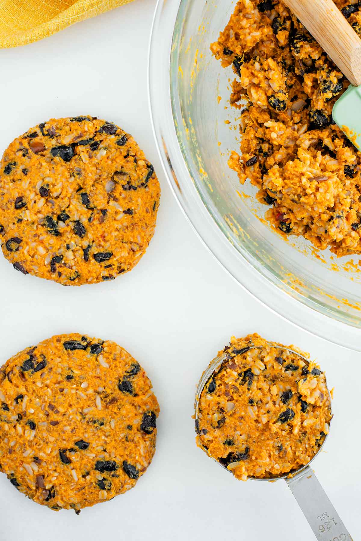 Top down view of sweet potato black bean burger patties being formed before cooking. Formed patties rest beside a measuring cup holding the burger mixture. The mixture and patties are golden in colour with black beans speckled throughout.