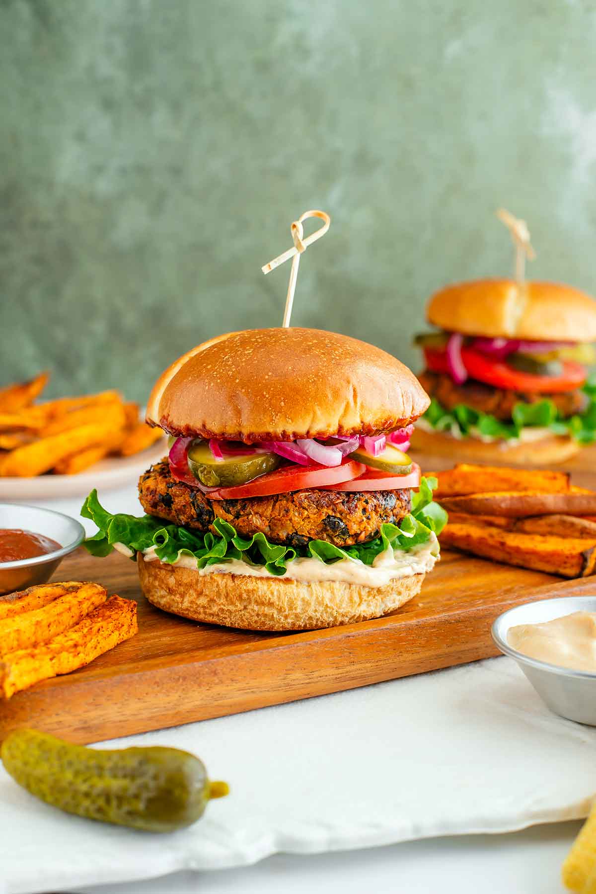 Side view of a vegan sweet potato black bean burger. The burger patty looks thick, crispy, and golden. The burger is loaded with chipotle mayo, lettuce, tomato, pickles, pickled red onion, and chipotle BBQ sauce. Another burger rests in the background.