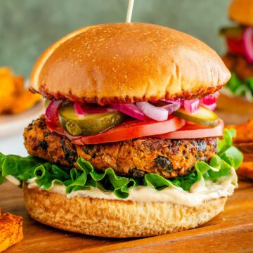 Side view of a vegan sweet potato black bean burger. The burger patty looks thick, crispy, and golden. The burger is loaded with chipotle mayo, lettuce, tomato, pickles, pickled red onion, and chipotle BBQ sauce.