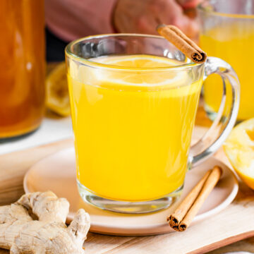Closeup side view of a mug of ginger turmeric tea. A woman pours hot water from a kettle into a second mug of tea in the background.