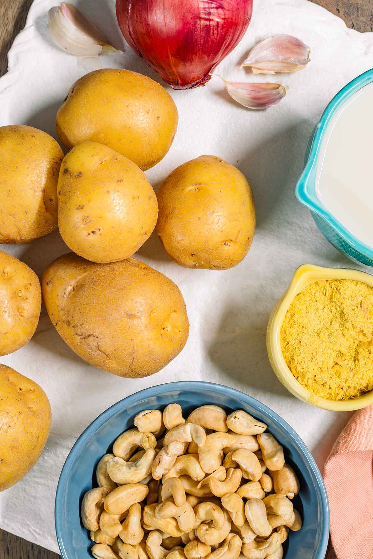 Top down view of ingredients for dairy free scalloped potatoes. Yellow potatoes are arranged next to small dishes of raw cashews, nutritional yeast and plant milk. A red onion and cloves of garlic rest at the top of the tray.