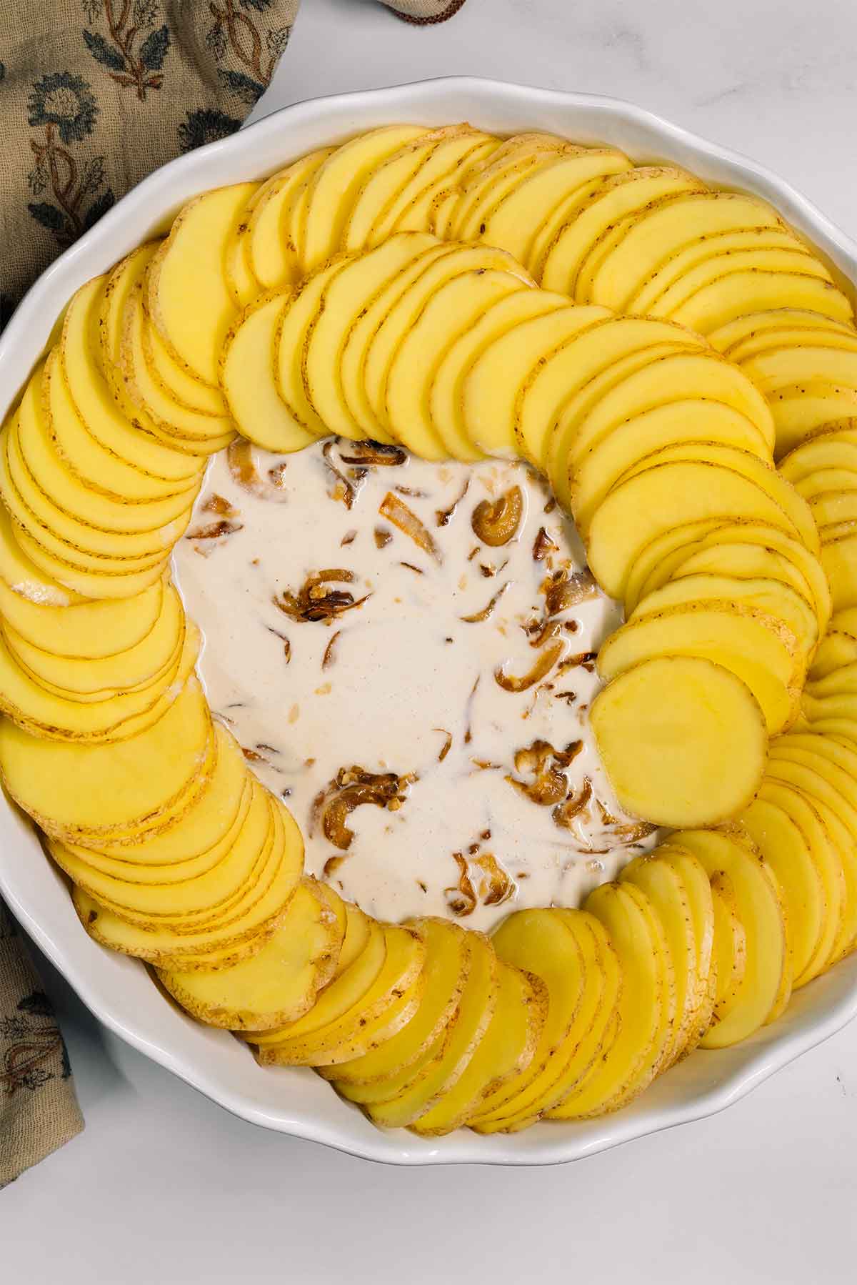 Top down process shot. Cream sauce and caramelized onions lay under thin slices of potatoes being arranged in a spiralled design inside a round baking dish.