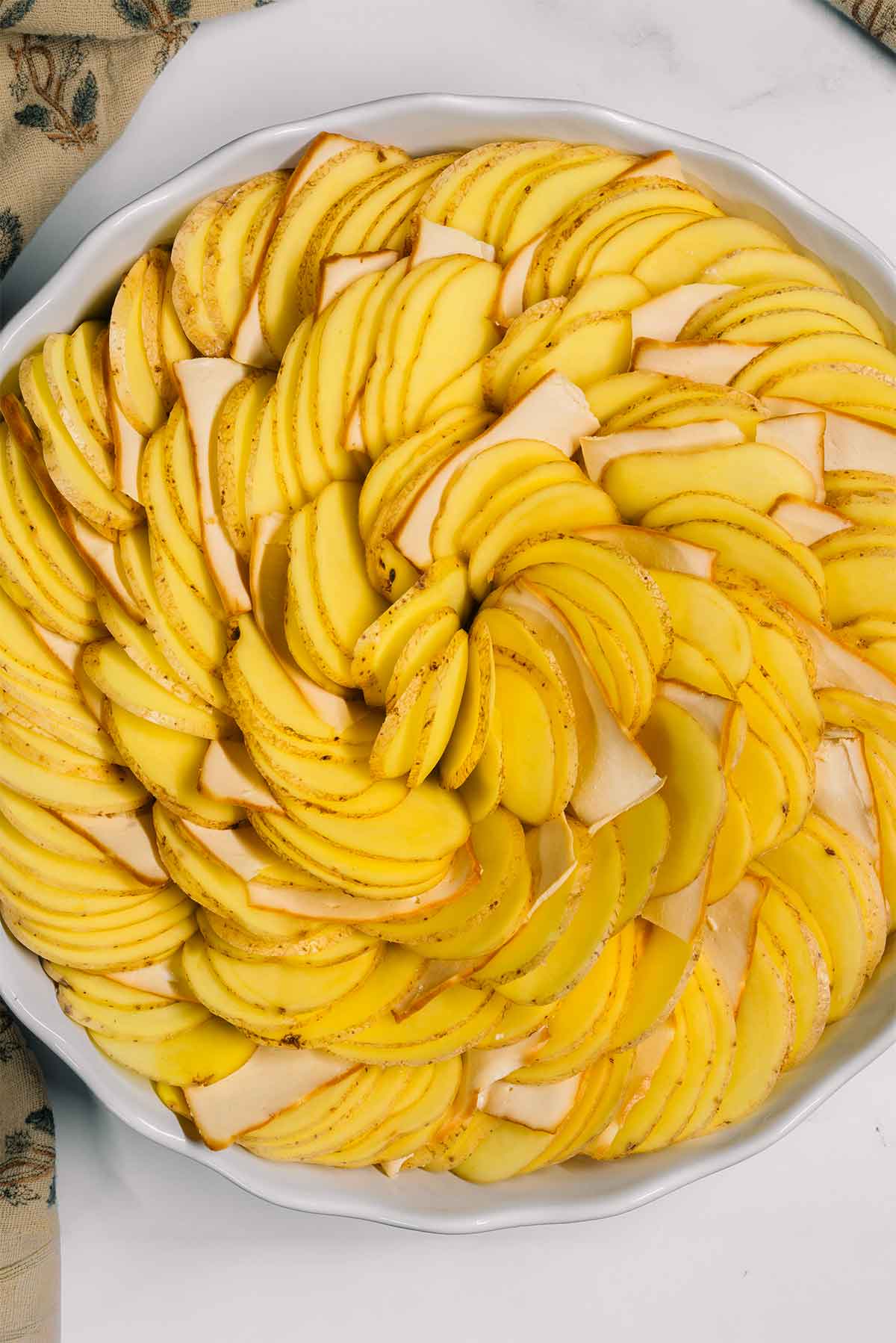 Top down process shot of sliced smoked tofu scattered between thin slices of potatoes. The potatoes are arranged in a spiralled design.