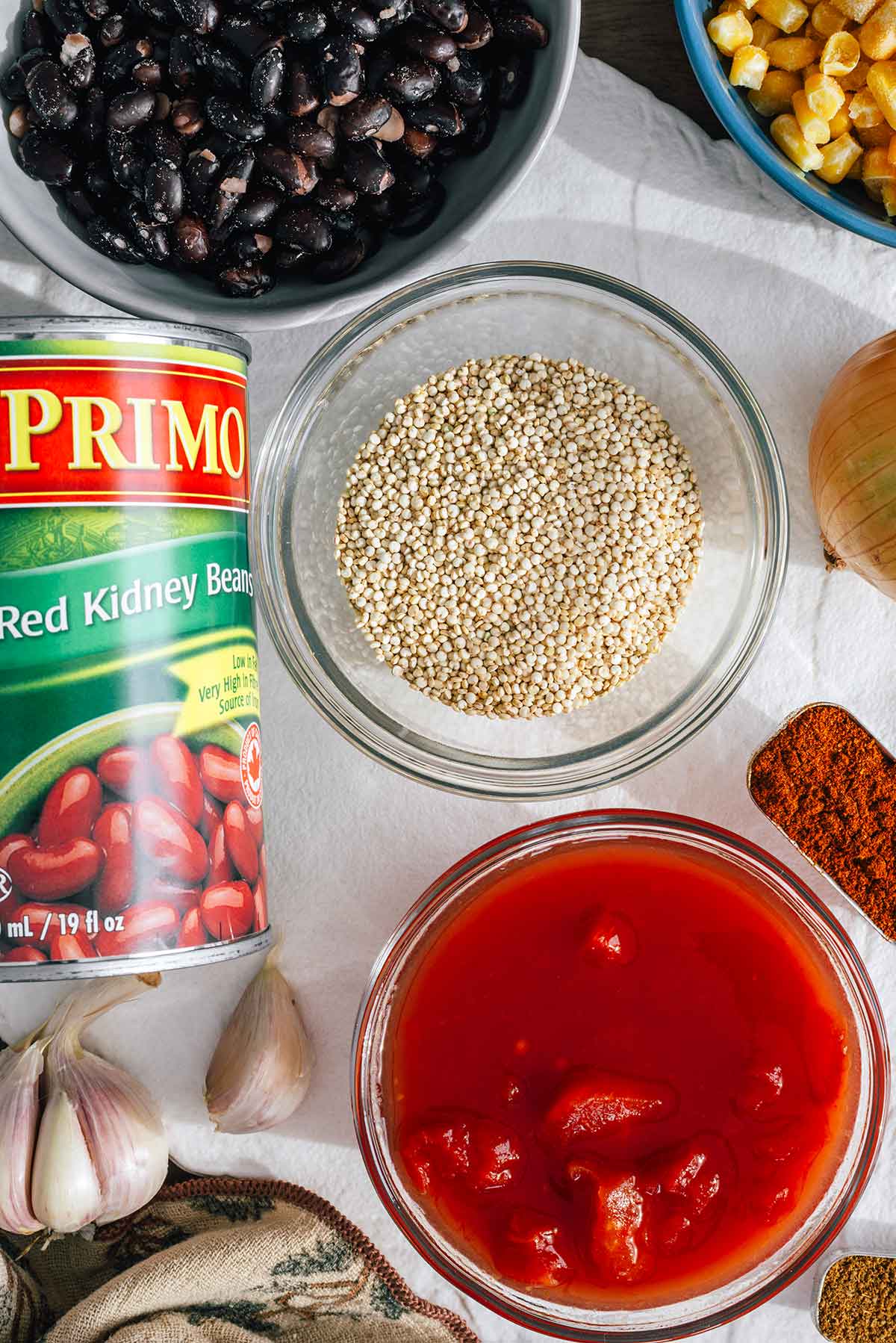 Top down view of ingredients on a tray. A small bowl of raw quinoa rests in the centre with kidney beans, black beans, corn, diced tomatoes, onion, garlic, chili powder, and cumin placed around.
