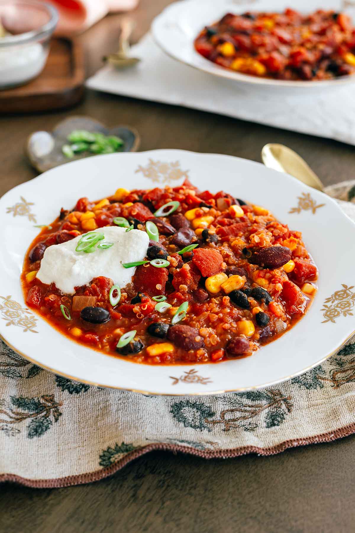 Side view of a vintage bowl filled with hearty quinoa chili. The chili is a chunky mixture of quinoa, kidney beans, black beans, and corn in a tomato sauce with a dollop of sour cream on top.