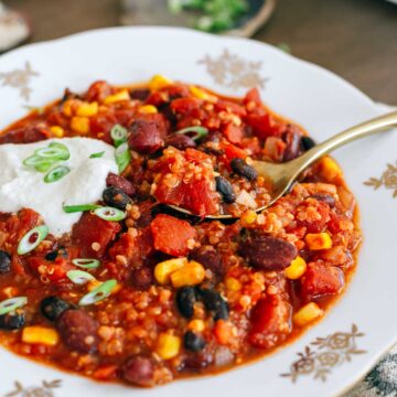 Side view of a spoon resting on top of a hearty bowl of chili. On the spoon, quinoa can be seen mixed with the tomato, bean, and corn stew. A dollop of sour cream is garnished with sliced green onion to finish of the look of the dish.