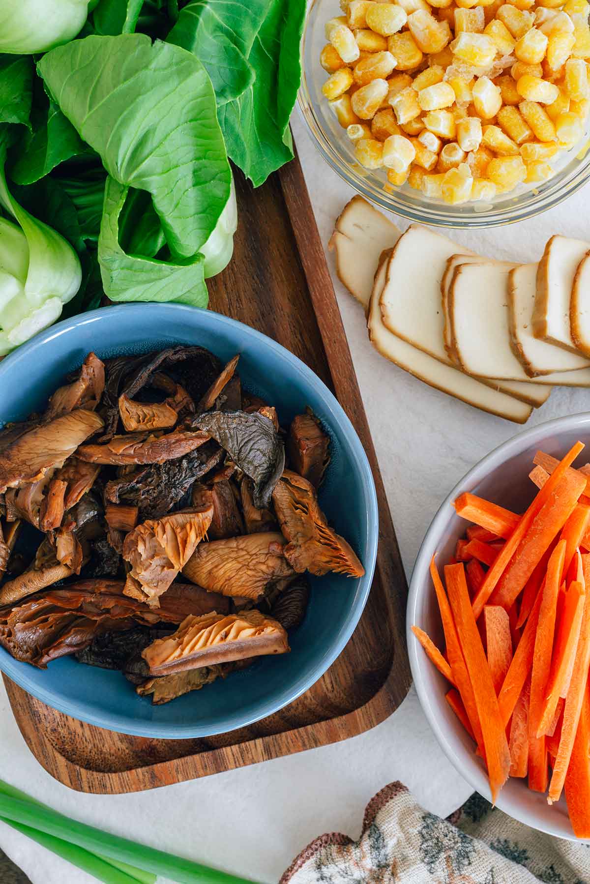 Top down view of ingredients for ramen toppings. A bowl of sliced gourmet mushrooms rests next to bok choy, corn, sliced smoked tofu, carrots and green onion.