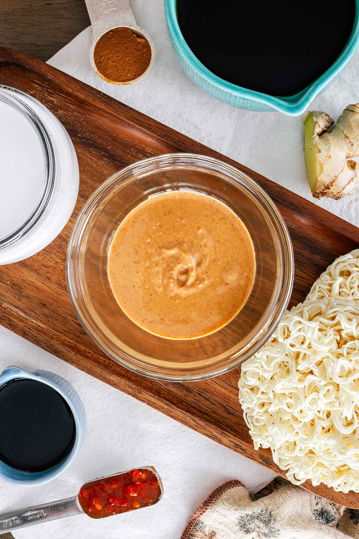 Top down view of ingredients for the broth. A small bowl of peanut butter is in the centre with coconut milk, tamari, miso paste, ginger, chili garlic sauce, and ramen noodle blocks positioned around.