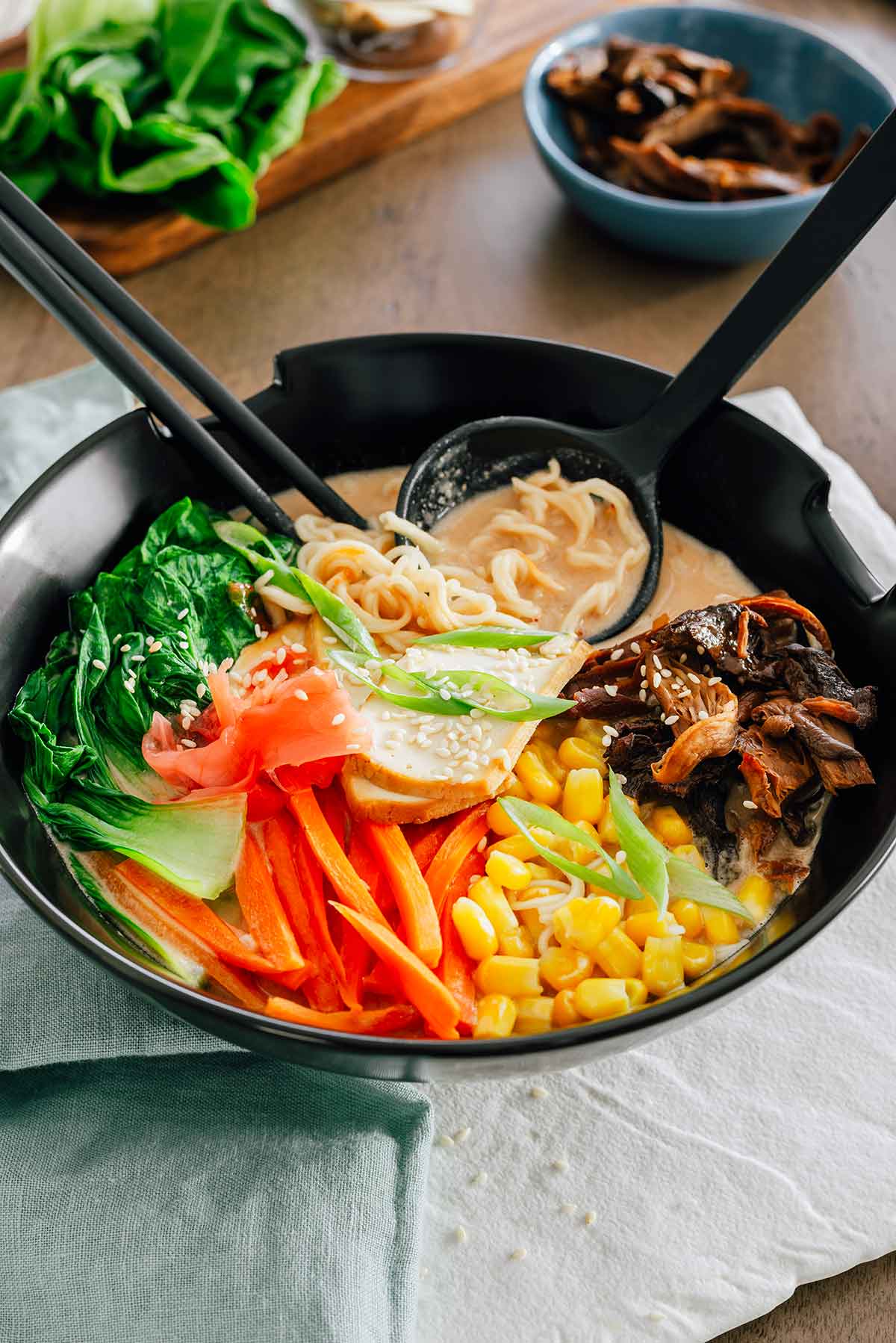 Side view of a colourful bowl of ramen sitting atop a white tray on a wooden table. A spoon rests in the bowl holding a creamy broth and curly ramen noodles. Sliced carrot, bok choy, mushrooms, corn, smoked tofu, green onion, and sesame seeds rest on top of the ramen noodles and broth.
