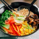Side view of a colourful bowl of ramen. A spoon rests in the bowl holding a creamy broth and curly ramen noodles. Sliced carrot, bok choy, mushrooms, corn, smoked tofu, green onion, and sesame seeds rest on top of the ramen noodles and broth.