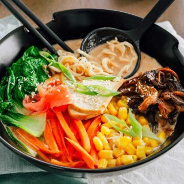 Side view of a colourful bowl of ramen. A spoon rests in the bowl holding a creamy broth and curly ramen noodles. Sliced carrot, bok choy, mushrooms, corn, smoked tofu, green onion, and sesame seeds rest on top of the ramen noodles and broth.