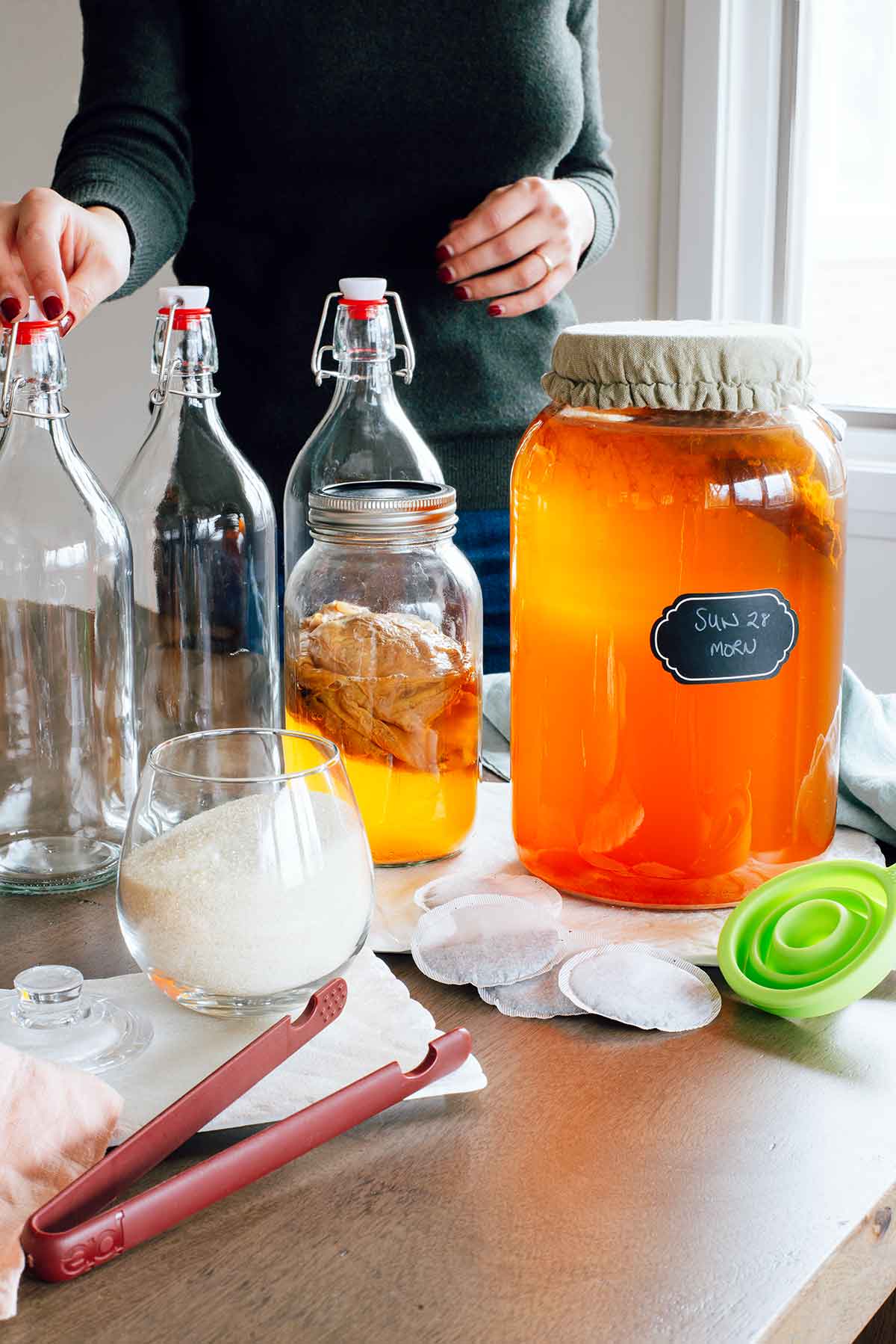 A 1 gallon glass brewing jar covered with a breathable cloth cover sits beside three 1 litre glass swing top bottles. A silicone funnel and silicone tongs are also visible amount the kombucha materials.