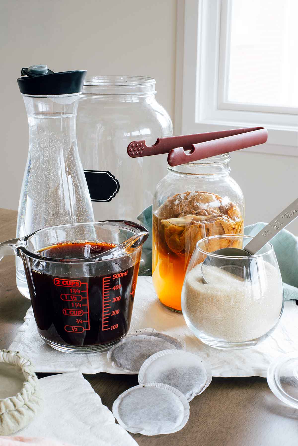 Ingredients for how to brew kombucha at home are displayed on a table. Brewed black tea, cane sugar and water surround a SCOBY pellicle and starter tea in a glass jar.