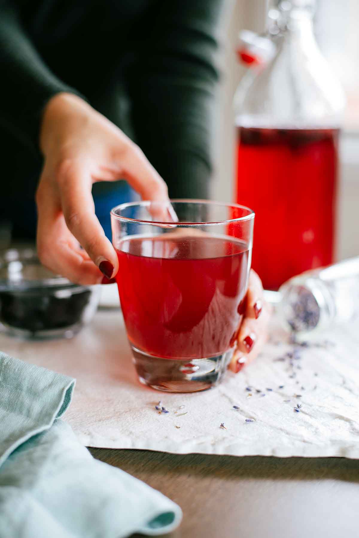 A hand reaches out for a glass of fizzy kombucha. The kombucha is a dark red colour and lavender buds are scattered on the tray around the glass. In the background is a glass bottle of kombucha with blueberries floating at the top.