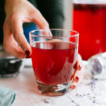 Side view of a hand reaching out for a glass of fizzy kombucha. The kombucha is a dark red colour and lavender buds are scattered on the tray around the glass. In the background is a glass bottle of kombucha with blueberries floating at the top.