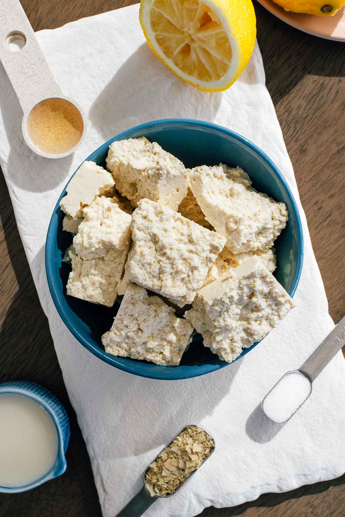 Top down view of ingredients in the sunlight. Crumbled tofu fills a small bowl surrounded by a halved lemon, soy milk, apple cider vinegar, salt, and nutritional yeast.