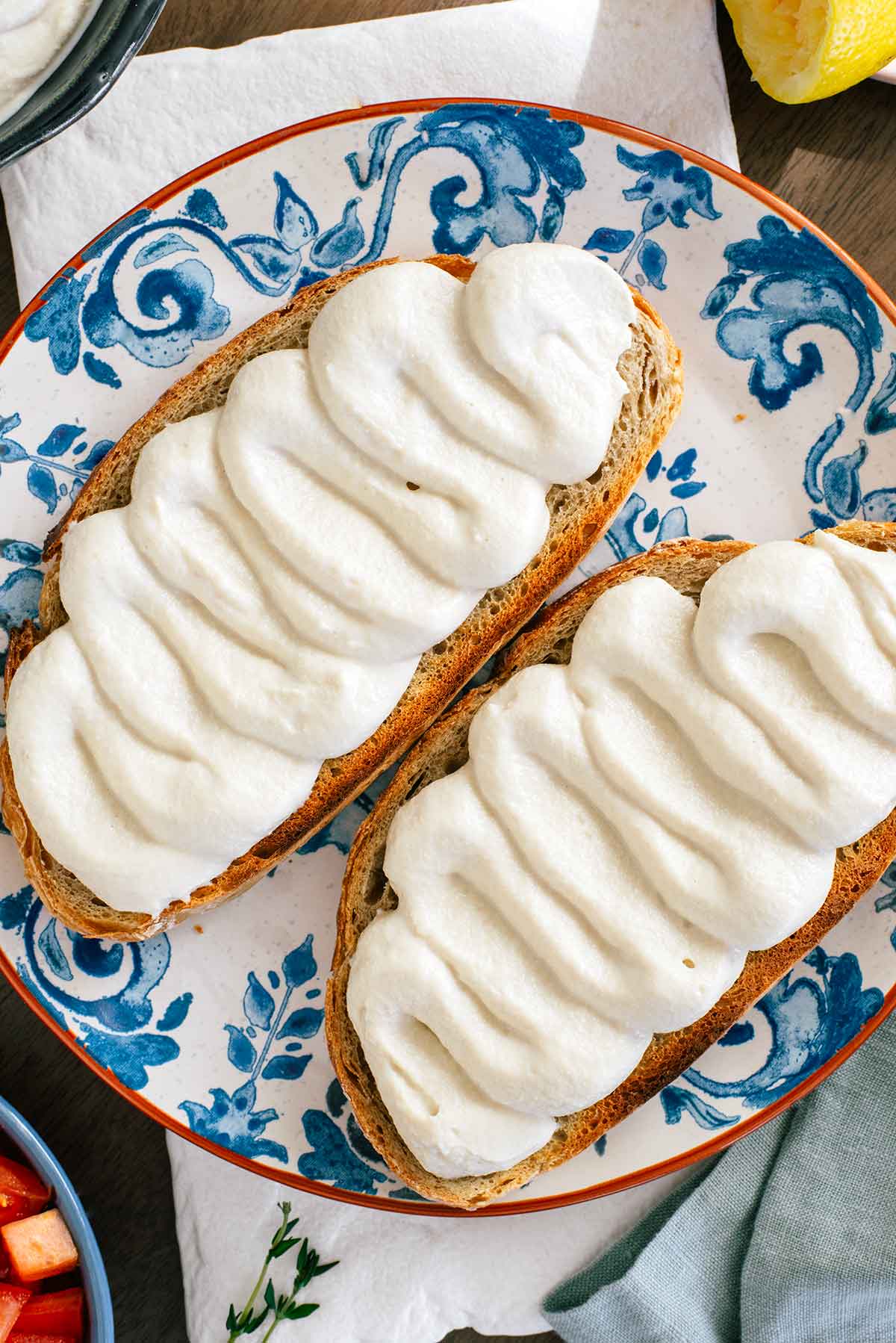 Top down view of fluffy whipped tofu piped in waves onto a slice of toasted bread. The bread sits atop a decorative clay plate and the tofu is creamy and pillowy. A lemon wedge, diced tomato, and fresh thyme surround the plate.