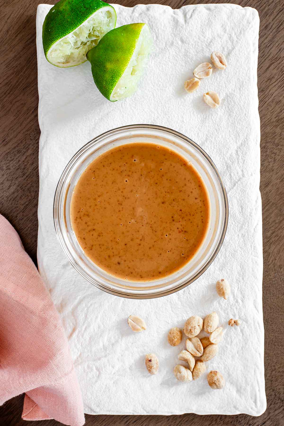 Top down view of a small bowl of peanut lime dressing atop a white tray. The dressing is thick and rich with squeezed lime wedges next to the bowl.