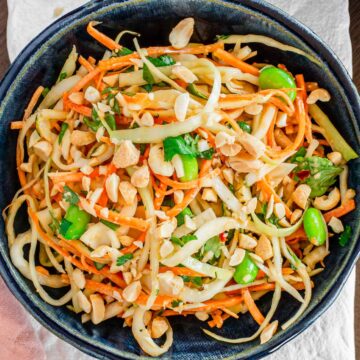 Top down view of a colourful cabbage salad in a small bowl. Green cabbage and carrot are shredded and mixed with bright green edamame and cilantro leaves. A peanut dressing lightly coats the salad which is topped with chopped roasted peanuts.