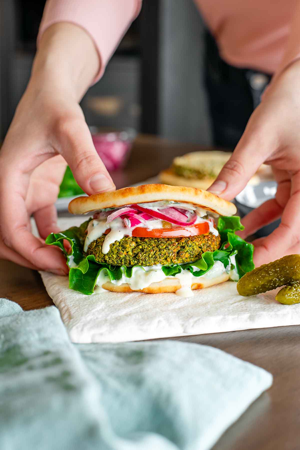 Side view of hands lifting a falafel burger from a tray. The burger patty is green and crispy on the outside. Creamy vegan tzatziki runs down the burger patty which is garnished with lettuce, tomato, pickles, and red onion.