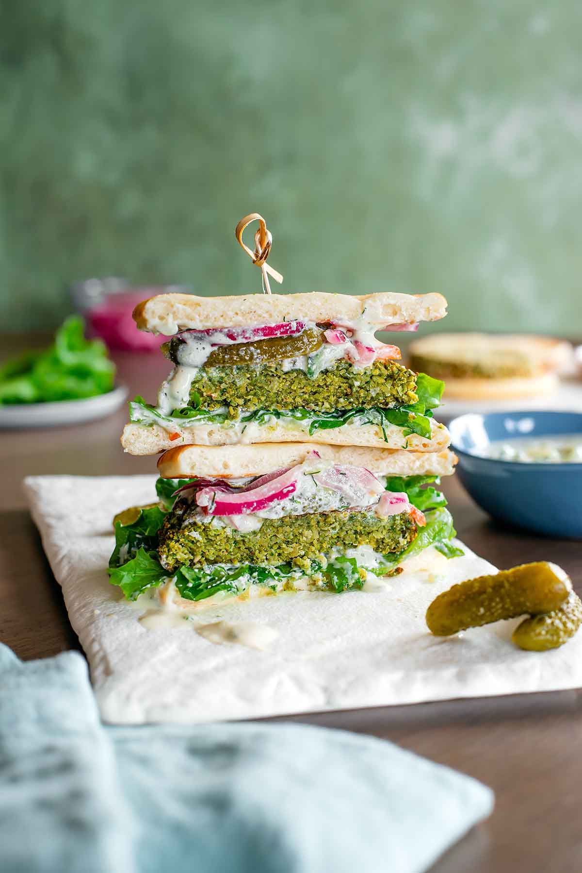 Side view of two halves of a falafel burger stacked on top of one another to show the fluffy green interior of the burger. The burger is loaded with a creamy vegan tzatziki sauce, lettuce, tomato, pickles and pickled red onion. Pita rounds replace a traditional burger bun.