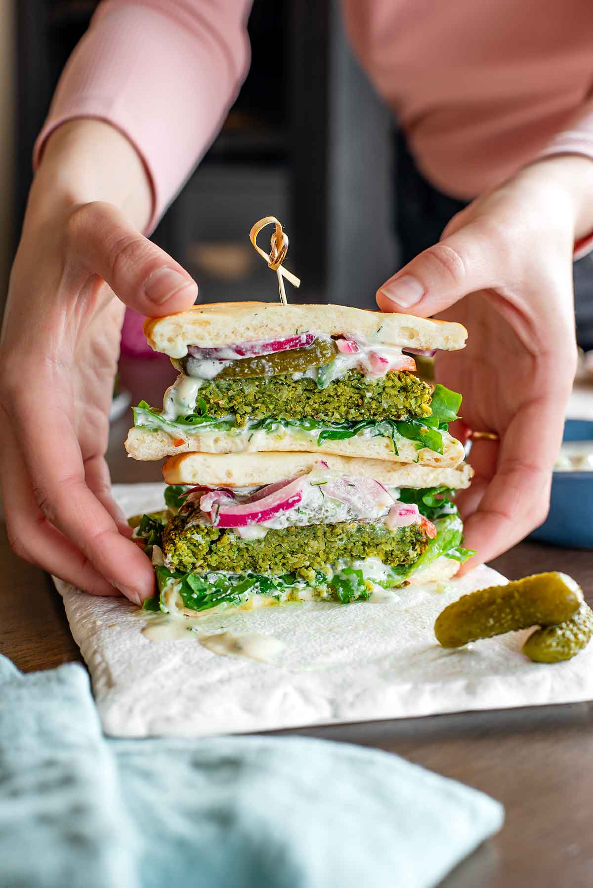 Side view of hands grabbing two stacked halves of a falafel burger. The burger is held together with a bamboo pick and green on the inside. The patty looks thick, fluffy inside, and crispy outside.