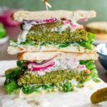 Two halves of a falafel burger are stacked showing the fluffy green interior of the falafel burger. The burger is loaded with a creamy vegan tzatziki sauce, lettuce, tomato, pickles and pickled red onion. Pita rounds replace a traditional burger bun.