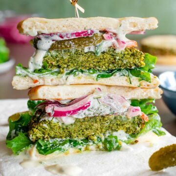 Two halves of a falafel burger are stacked showing the fluffy green interior of the falafel burger. The burger is loaded with a creamy vegan tzatziki sauce, lettuce, tomato, pickles and pickled red onion. Pita rounds replace a traditional burger bun.