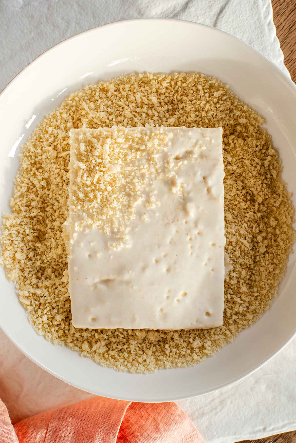 Top down view of a rectangular slice of tofu being covered in panko breadcrumbs. The tofu has been dipped in a flour and cornstarch batter to help the breadcrumbs adhere.