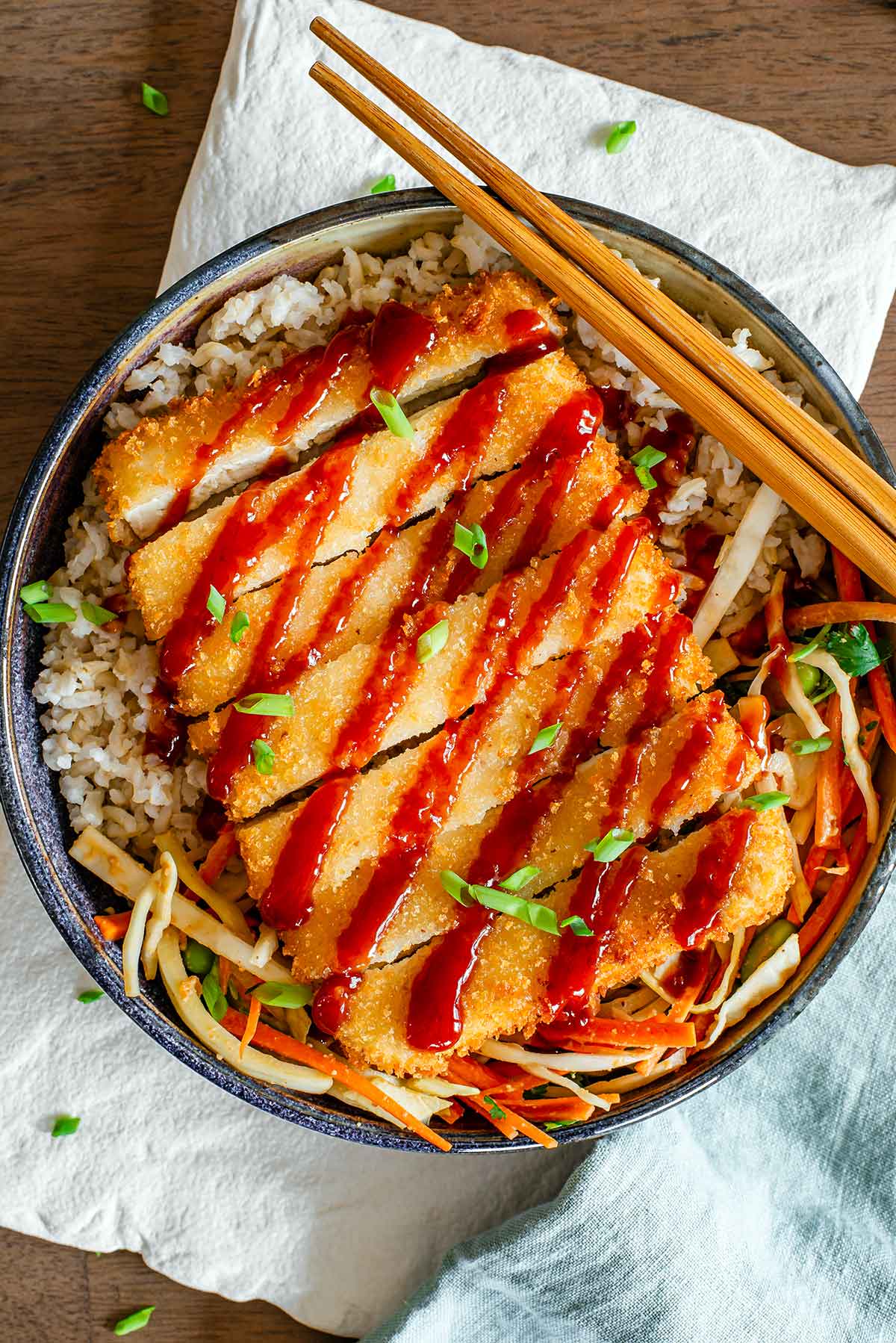 Top down view of tofu katsu atop a bed of brown rice and crunchy cabbage salad. The katsu is breaded and golden with a deep red katsu sauce drizzled on top.