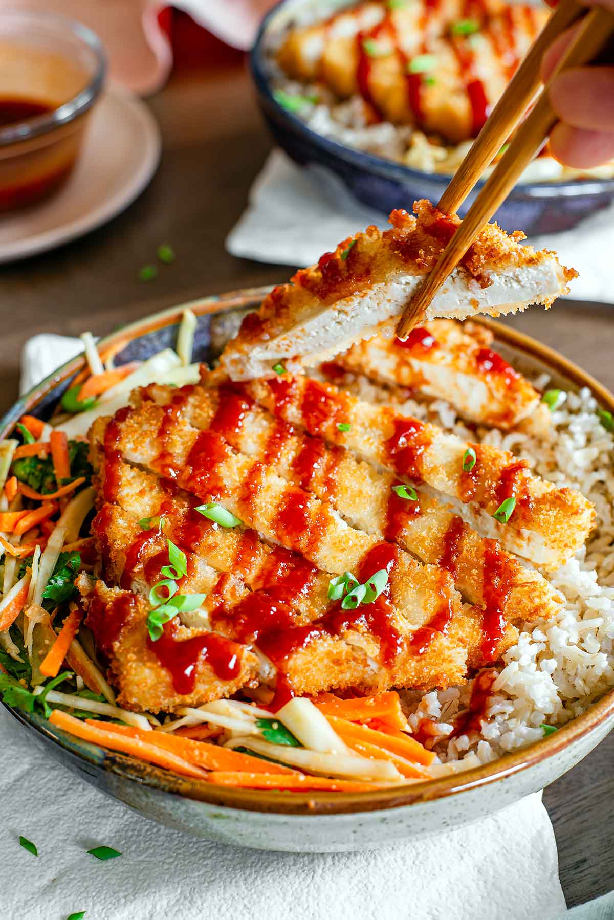 Side view of chopsticks lifting a slice of crispy tofu katsu from a bowl. The tofu looks juicy on the inside and has a golden breading on the outside. The remainder of the katsu rests on a bed of brown rice and cabbage salad and is drizzled with a deep red katsu sauce. Extra sauce rests in the background.