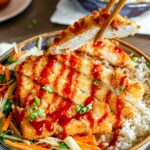 Side view of chopsticks lifting a slice of crispy tofu katsu from a bowl. The tofu looks juicy on the inside and has a golden breading on the outside. The remainder of the katsu rests on a bed of brown rice and cabbage salad and is drizzled with a deep red katsu sauce.