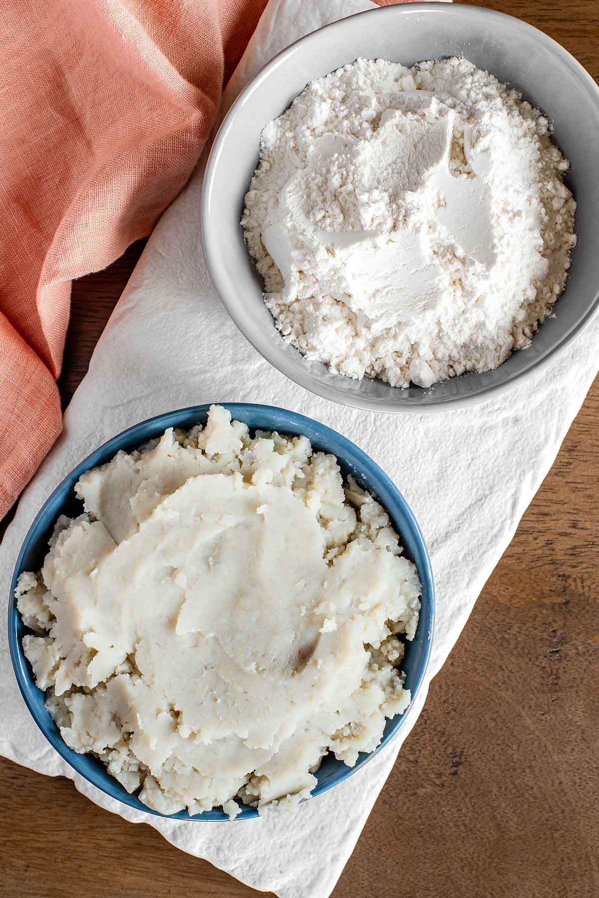 Top down view of ingredients for mashed potato gnocchi. Leftover mashed potatoes fill a small bowl resting next to another small bowl of all-purpose flour.