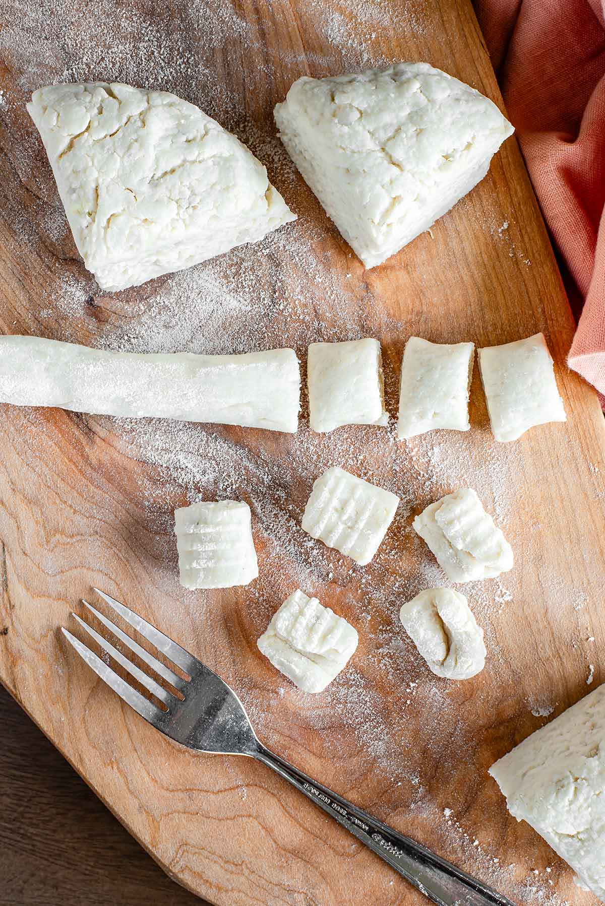 Top down view of the gnocchi dough in different stages. Portions of the larger dough ball rest beside a portion rolled out into a tube and partially cut into smaller pieces of gnocchi. Finally, there are pieces of gnocchi that have been rolled with a nearby fork.