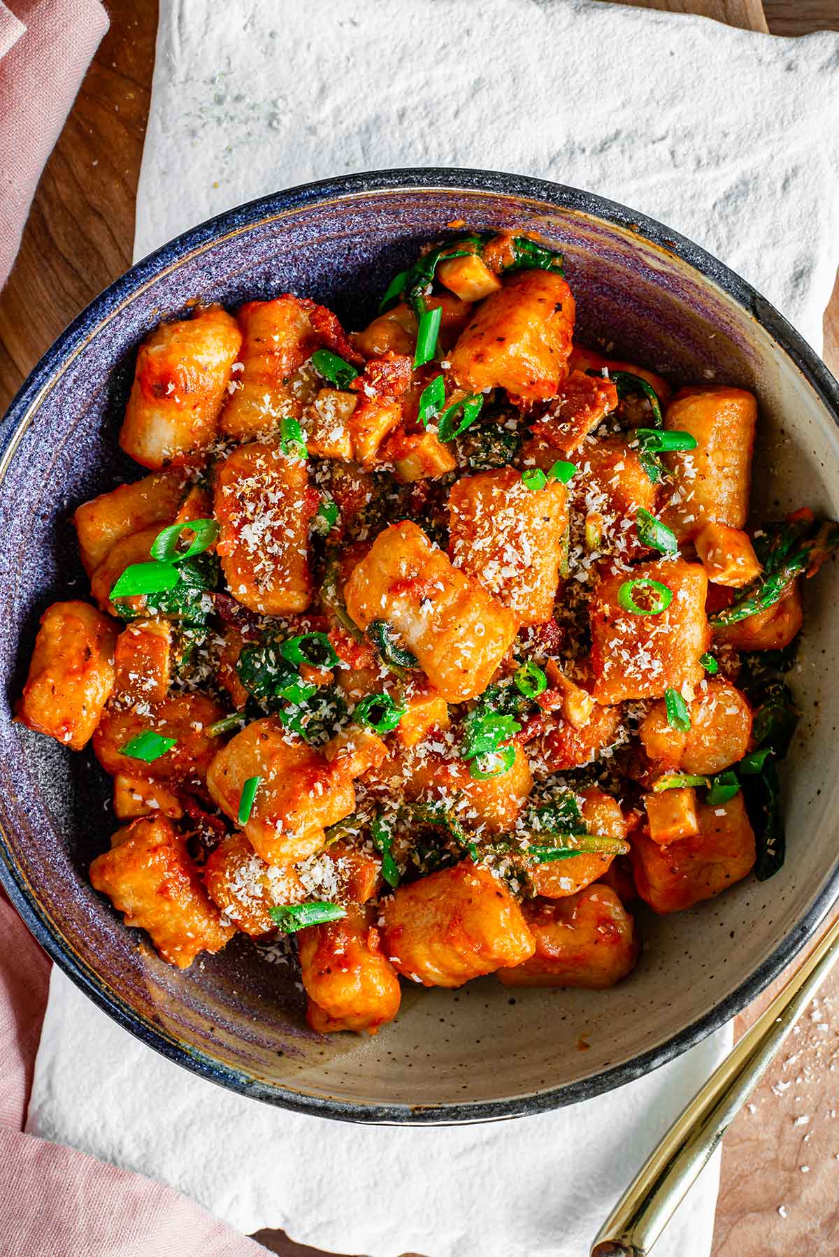 Top down view of a bowl of fluffy gnocchi made from leftover mashed potatoes. The gnocchi is ribbed and covered in a creamy tomato sauce with sun-dried tomatoes, smoked tofu, spinach, green onion, and vegan parmesan cheese.