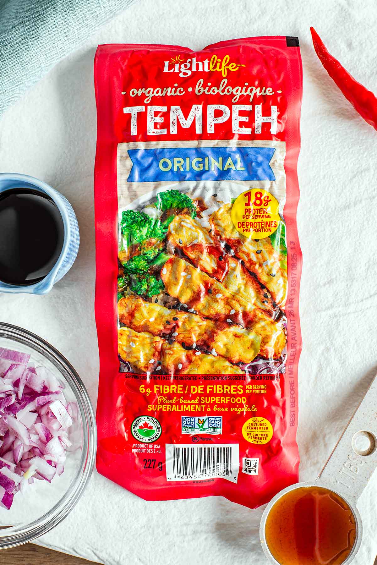 Top down view of a store-bought package of tempeh surrounded by diced red onion, a red chili pepper, tamari, and maple syrup.