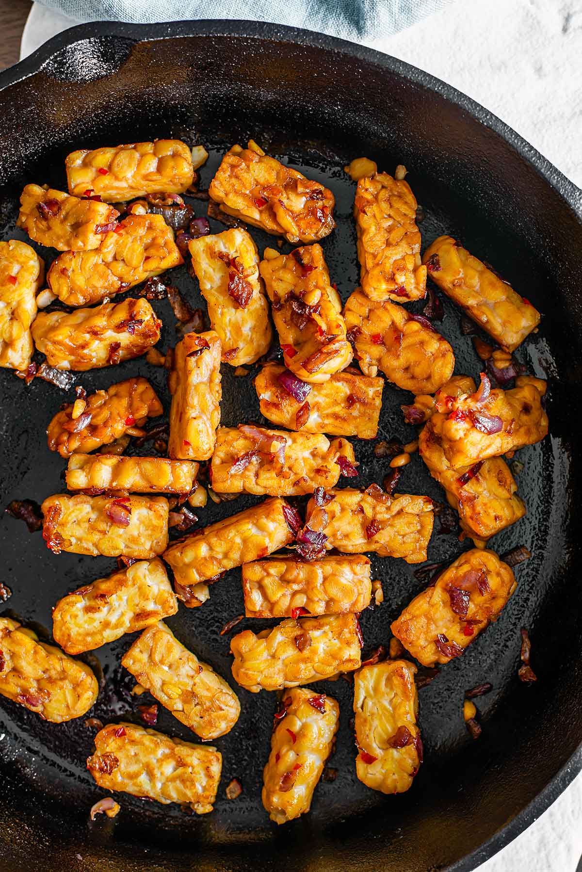 Top down view of golden brown tempeh in a cast iron frying pan. The tempeh is speckled with sautéed red onion and minced chili pepper.